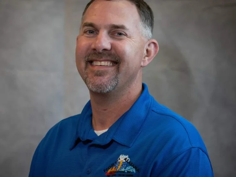 Smiling man with short hair and goatee wearing a blue collared shirt against a gray background.